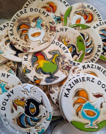 Zestaw okrągłych, ręcznie malowanych ceramicznych magnesów, z których jeden przedstawia kolorowego koguta, z widocznym napisem 'KAZIMIERZ DOLNY'.
