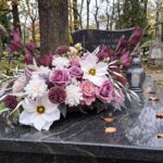 Elegancka wiązanka nagrobna w odcieniach fioletu, różu i bieli, leżąca na ciemnym, granitowym nagrobku. Obok kompozycji kwiatowej stoi szklany znicz na tle jesiennego cmentarza.