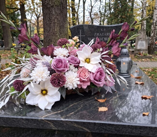 Elegancka wiązanka nagrobna w odcieniach fioletu, różu i bieli, leżąca na ciemnym, granitowym nagrobku. Obok kompozycji kwiatowej stoi szklany znicz na tle jesiennego cmentarza.